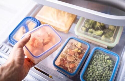 Person places a blue-lidded container into a freezer among other meal-prep containers with broccoli and peas nearby.