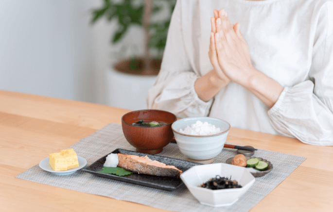 Person with hands in prayer before a Japanese-style meal set on a tray: grilled salmon, rice, miso soup, tamagoyaki, pickles and seaweed.