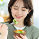 Young woman smiles while holding a wooden bowl of salad and about to take a bite with a fork.