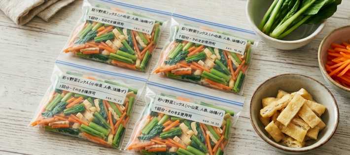Four clear resealable bags of chopped vegetable mix (green beans, carrots, bamboo shoots) on a light wood table, with bowls of greens and fried tofu nearby, ready for cooking.